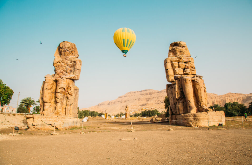 The Colossi of Memnon is two giant statues of Pharaoh Amenhotep III. Between the statues, a yellow hot air balloon rises in the clear blue sky.
