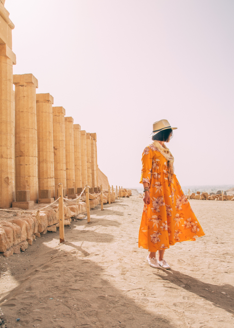 A woman wearing a long orange dress with an all-over floral pattern poses facing away from the camera. She is in Egypt, standing on sandy ground next to ancient building ruins with tall stone pillars.