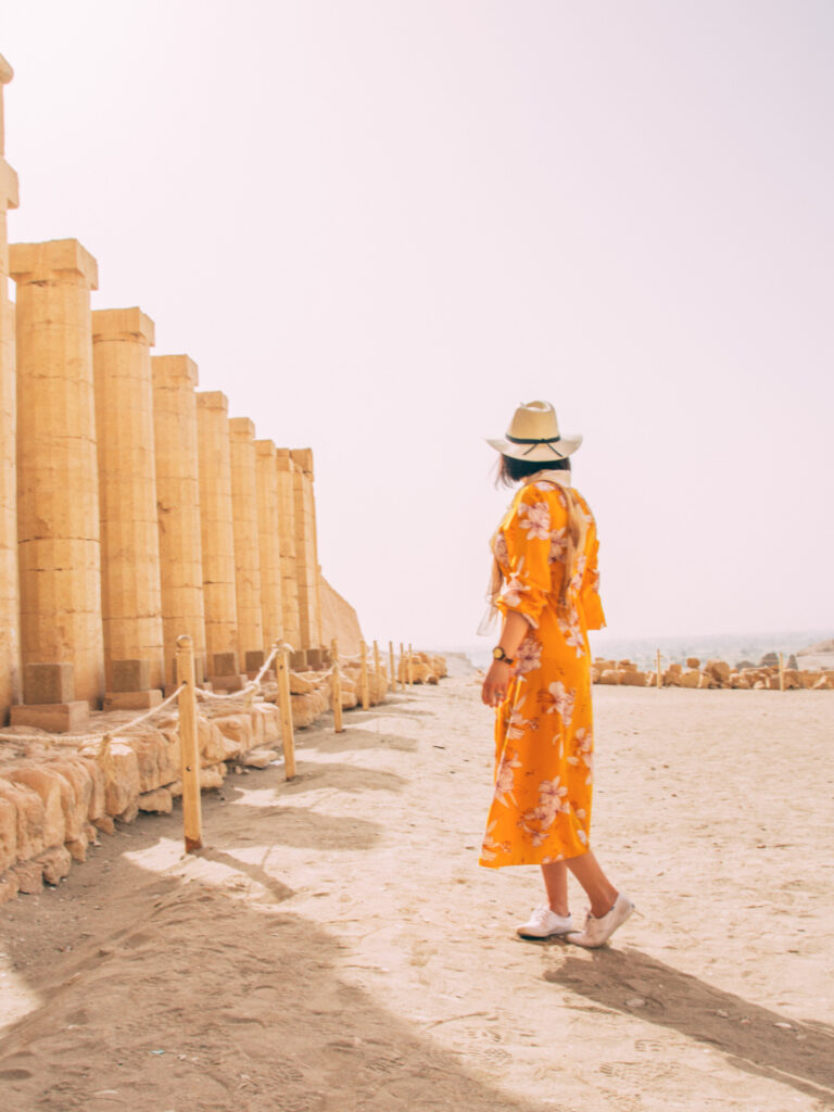A woman wearing an orange floral dress poses with her back facing the camera. She's wearing a hat and white shoes, and stands on sandy ground next to ancient ruins with tall stone pillars.