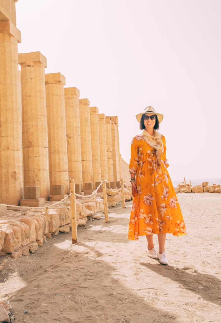 A woman with short brown hair wears a long orange dress with a floral pattern, linen scarf, hat, and sunglasses. She poses next to tall stone pillars that are part of ancient ruins in Luxor, Egypt.