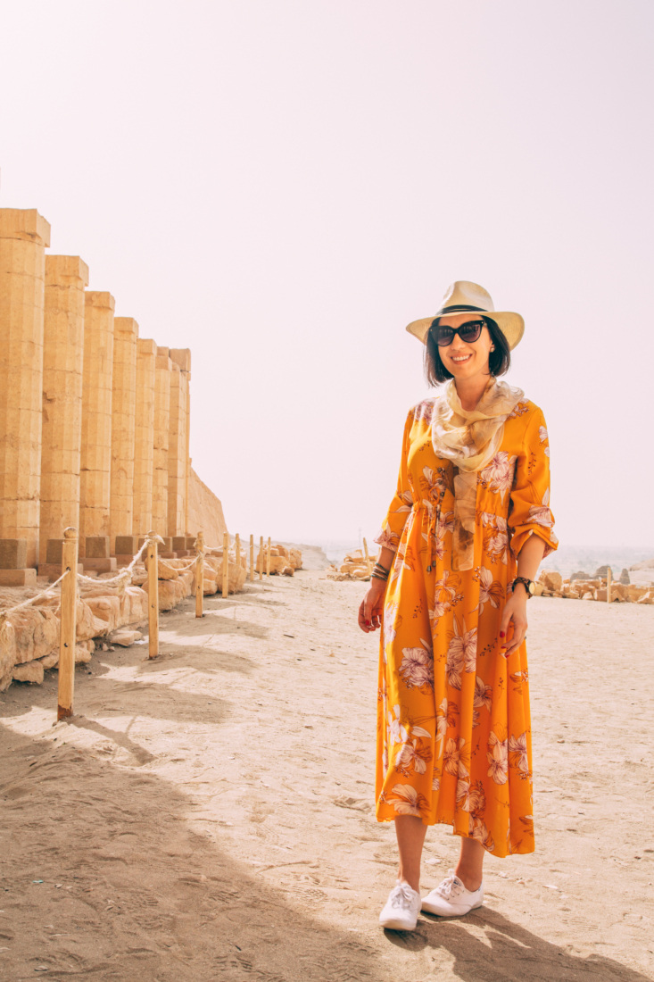 A woman poses next to tall stone pillars in Luxor, Egypt. She's wearing an orange dress with a floral pattern, hat and sunglasses, and is smiling at the camera.
