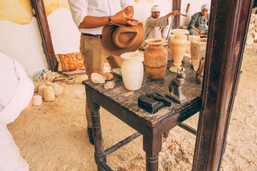 A man with a hat in his hand observing alabaster vases in Egypt with people sitting on the ground in the background. 