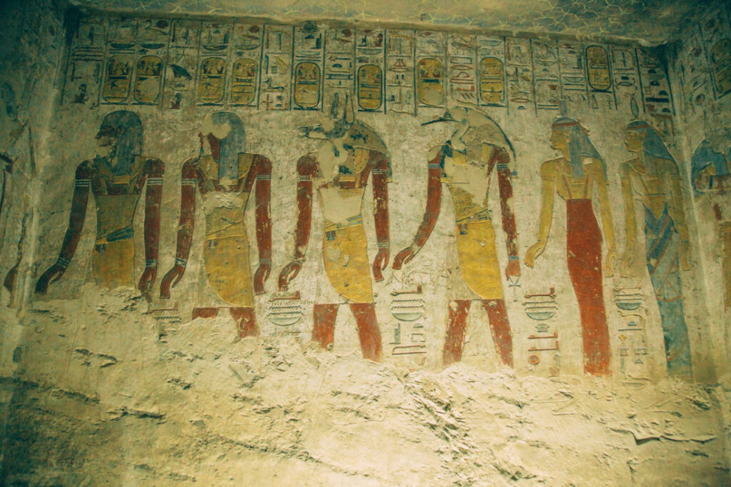 The remnants of ancient Egyptian murals on a stone wall inside a tomb at the Valley of the Kings. The art and hieroglyphs crumble away, exposing bare stone at the bottom of the image.
