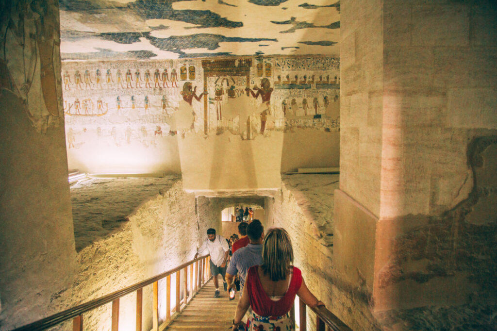 Tourists file down a walkway inside a tomb in the Valley of the Kings. There are well-lit alcoves and pillars on either side of the walkway, and the stone walls are covered in ancient artwork.