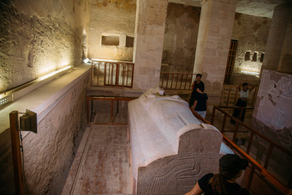 Tourists mill around a giant stone sarcophagus inside a tomb at the Valley of the Kings. The walls of the tomb is lit by spotlights, illuminating the art on the walls.