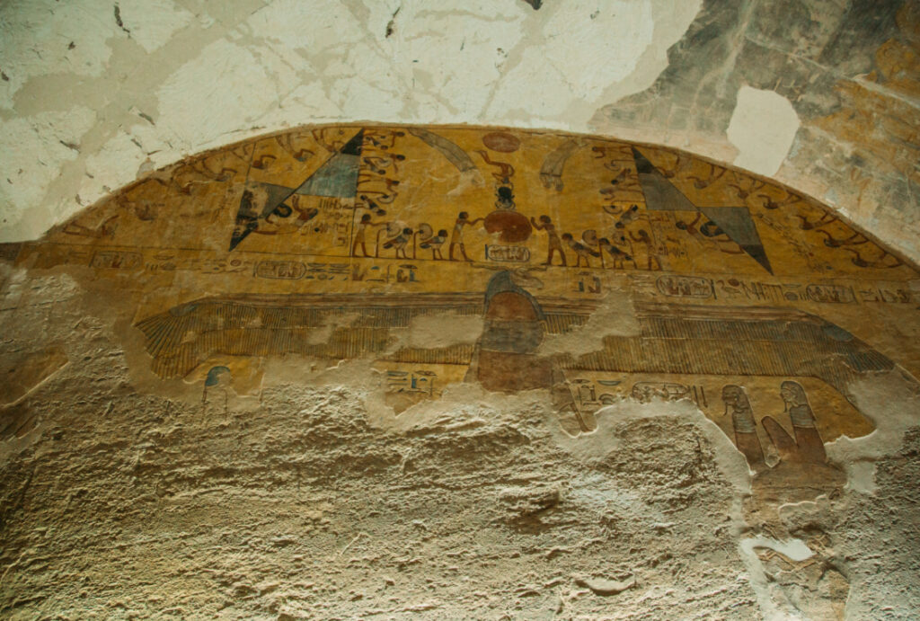 A partial mural remains at the top of an arched ceiling inside a tomb at the Valley of the Kings. The bottom of the mural crumbles away, exposing the natural stone walls.