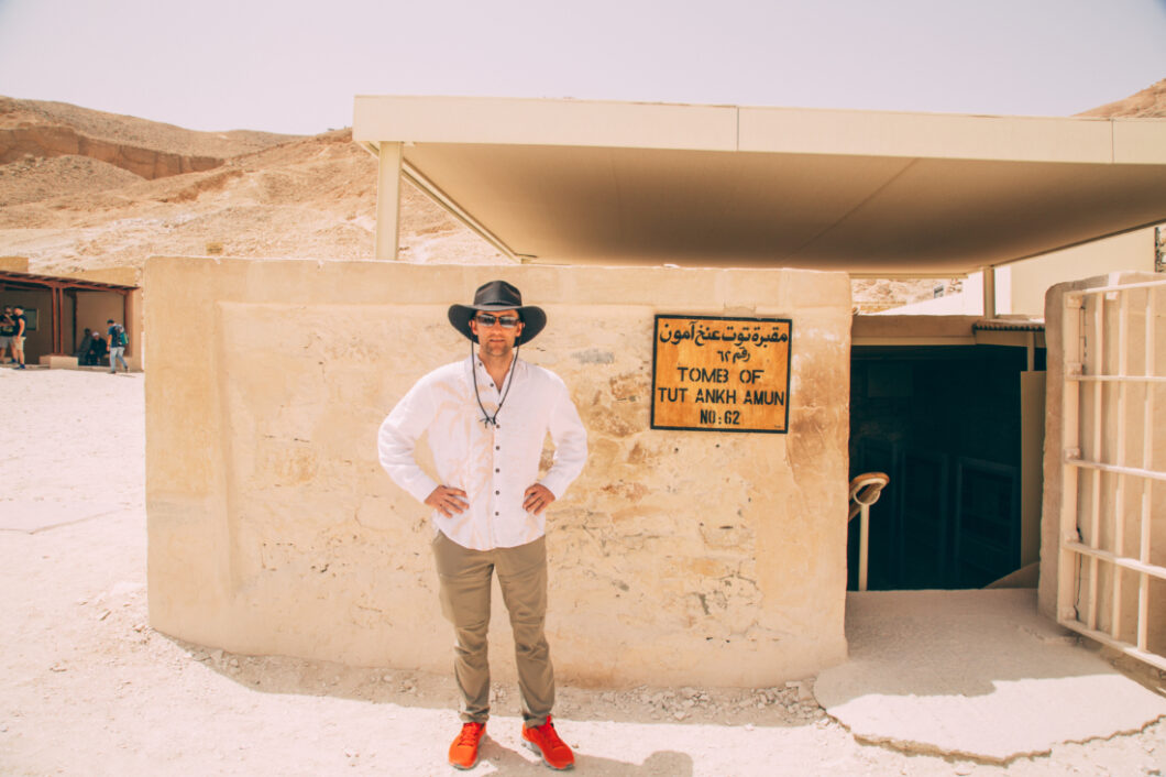 A man wearing a white long sleeve shirt, khaki pants, and a black hat poses with his hands on his hips. He's standing outside the entrance to the tomb of Tut Ankh Amun in Egypt.