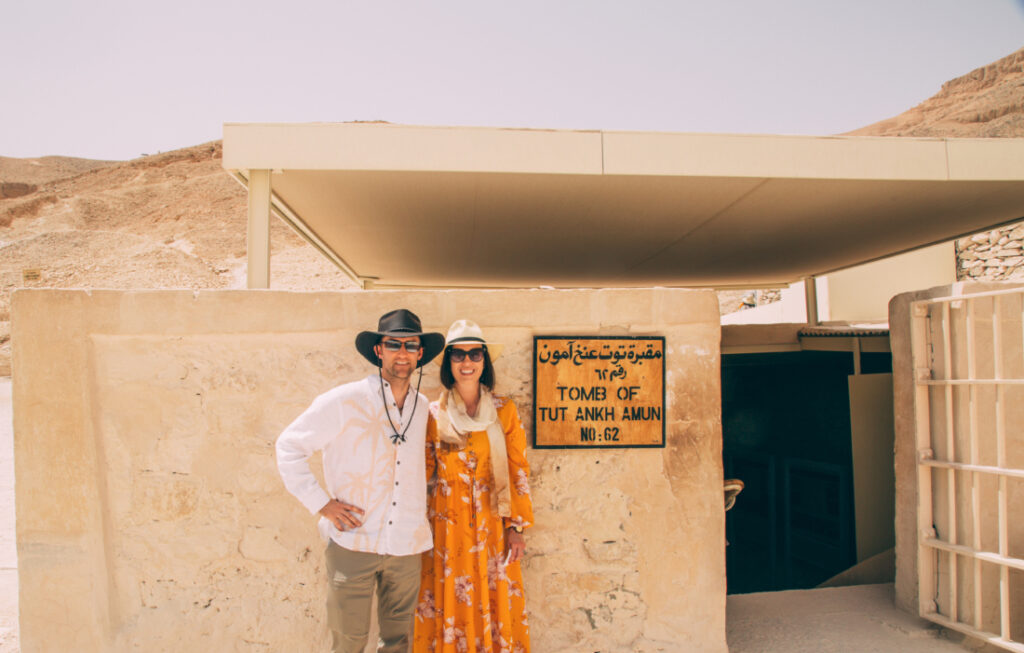 A man and woman wearing hats and long clothing, smiling and standing in front of the entrance to the Tomb of King Tut in Egypt