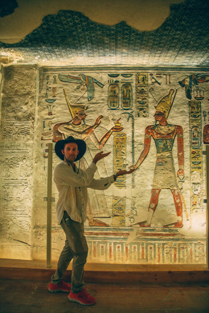 A man poses next to a wall filled with ancient artwork carvings of Egyptian Pharaohs. The man is mimicking the pose of the Pharoah. 
