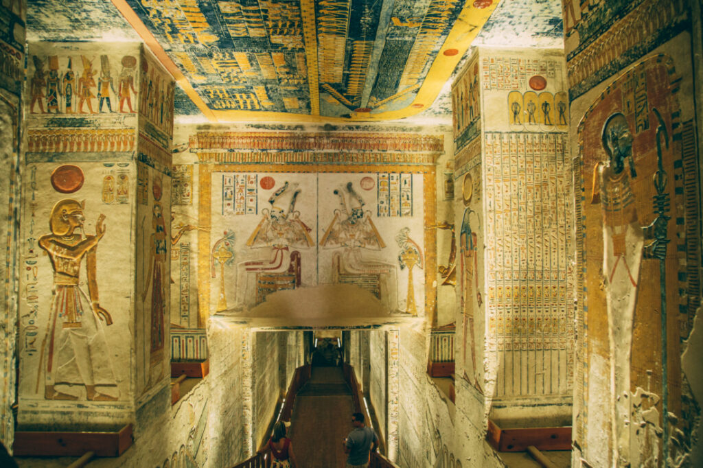 A hallway descends down into a tomb at the Valley of the Kings. The stone walls are covered with ancient artwork carvings that are well-preserved and still very colorful. 