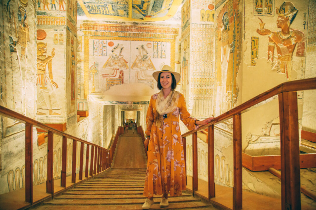 A woman stands on a wooden walkway inside a tomb in the Valley of the Kings. The stone walls of the tomb are covered with colorful carvings. The woman is wearing a long orange floral dress, a scarf, and a sunhat.