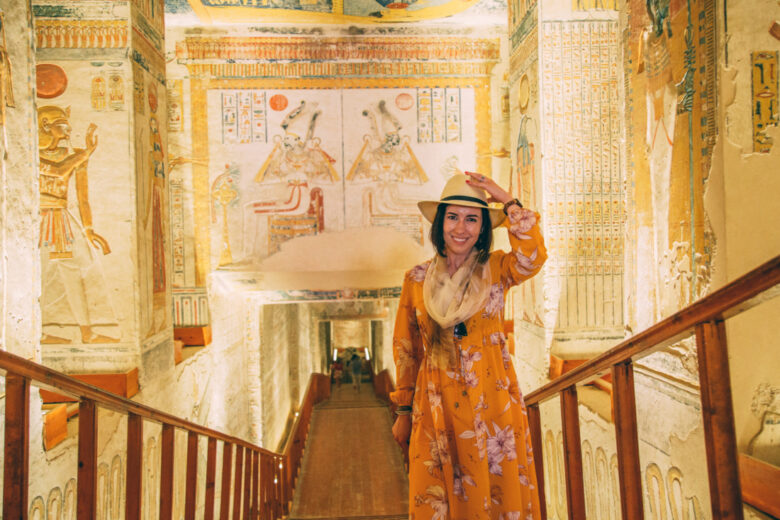 A woman stands inside the entry way of a tomb in Egypt. The walls are covered in ancient carvings and artwork. She's wearing a yellow floral dress, scarf, and a hat.
