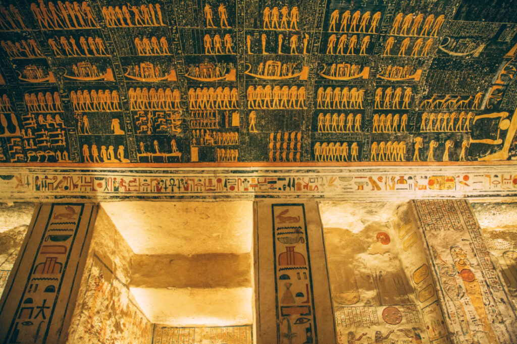 An image showing the ceiling and wall carvings inside the tomb of King Ramses V. The ceiling is painted a dark color, and the walls are a natural stone color.
