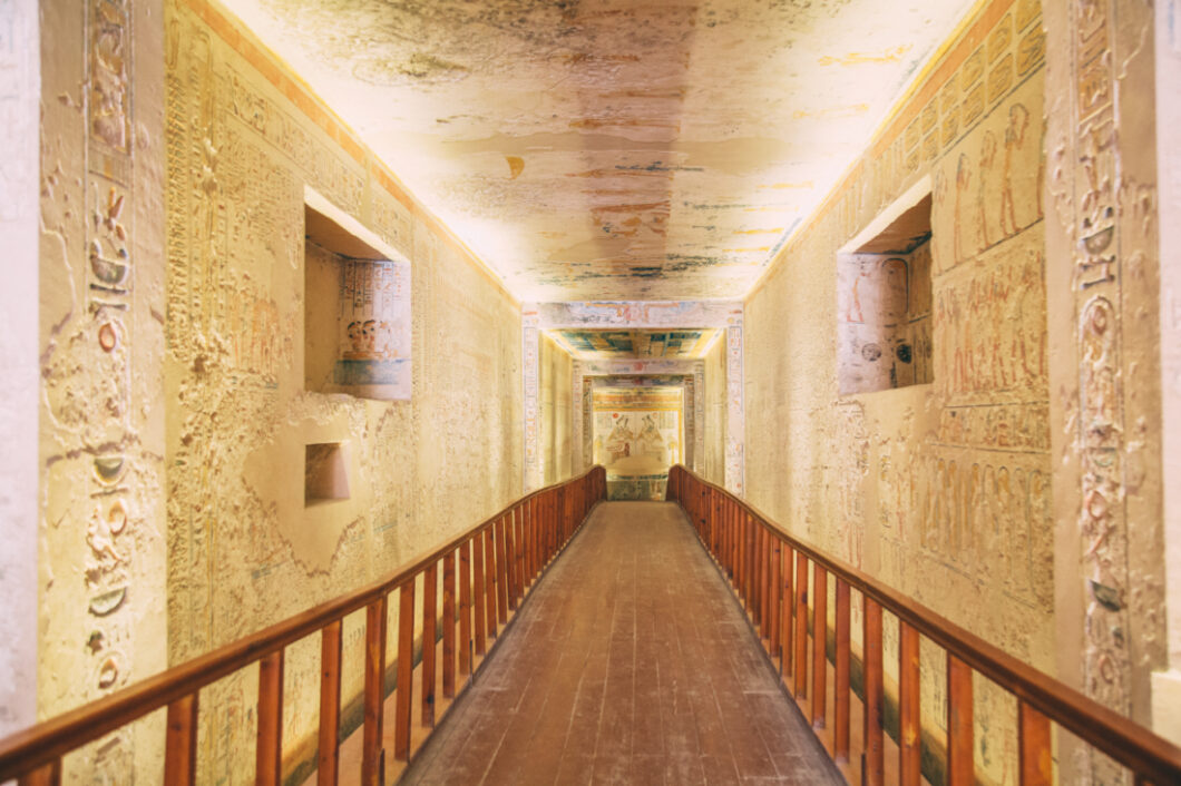 A long hallways in the tomb of King Ramses V. The stone walls are lined with cared hieroglyphs and artwork and there's a long wooden walkway.