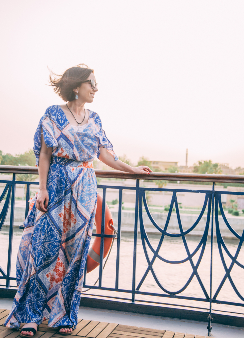 A woman stands on the deck of a Nile River cruise ship, looking out at the landscape. She's wearing a floor-length dress with a blue boho floral pattern that's accented with shades of light blue, tan and peach. She is wearing sunglasses, and her short hair is blowing in the wind.