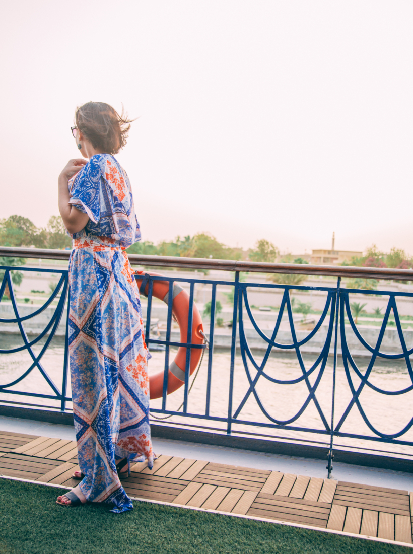 A woman stands on the deck of a river cruise ship with her back to the camera. She's wearing a long maxi-length blue floral print dress.