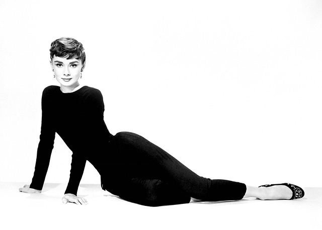 Black and white photo of Audrey Hepburn laying down propped up with her hands, wearing a black dress and black heels with short hair.