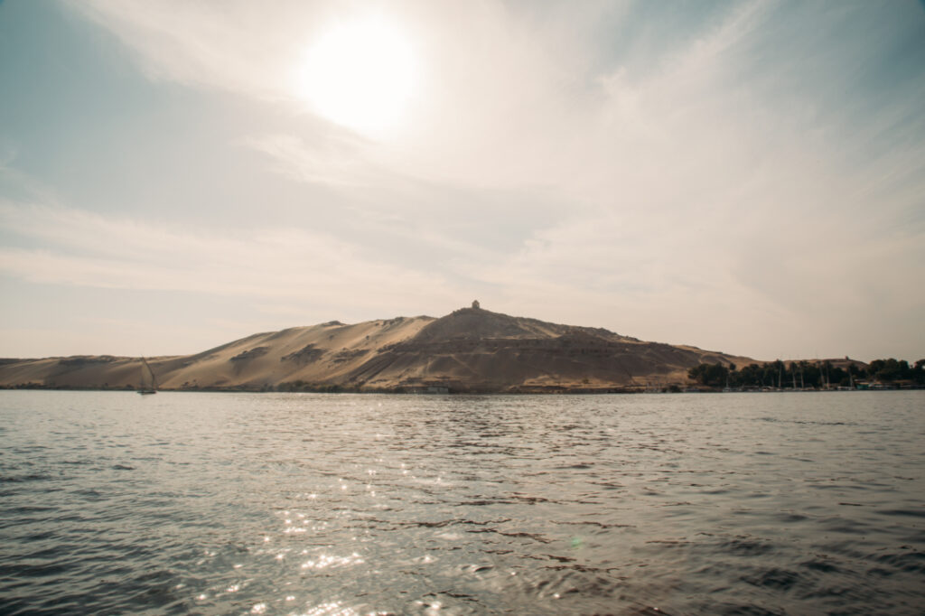 A large, sandy mountain stands tall against a clear, sunny sky on the riverbank of the Nile river near Aswan.