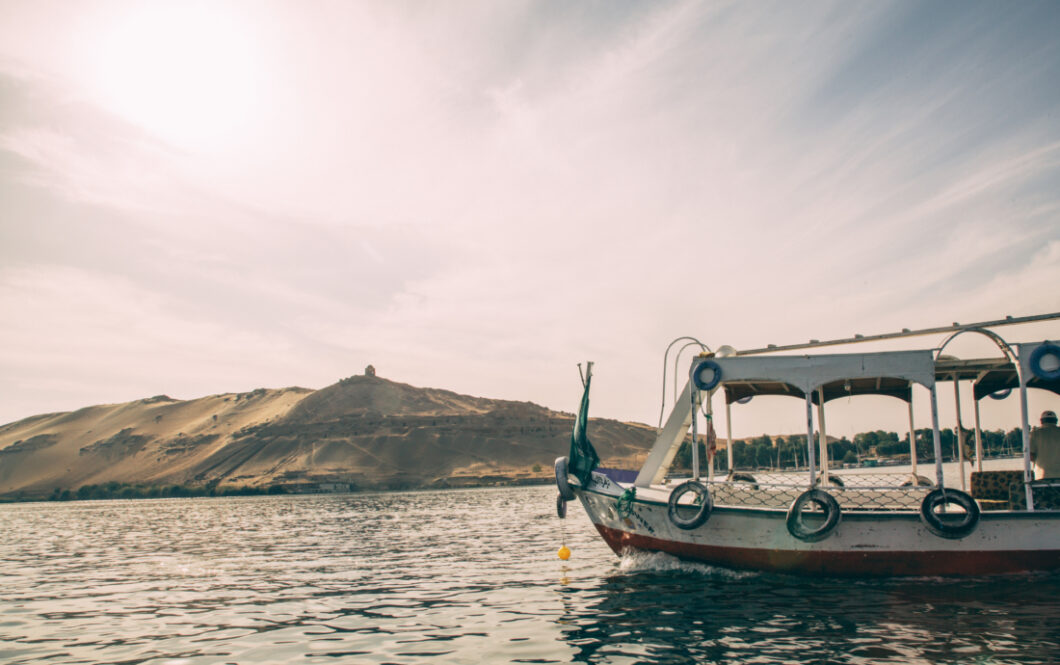 A river boat travels along the Nile River outside of the Nubian Village in Aswan. In the background are large sandy mountains and hills making up the riverbank landscape.