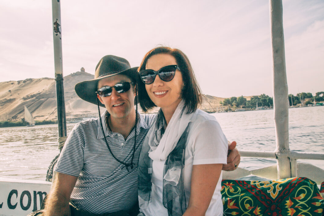 A man and woman couple pose with their arms around each other, smiling at the camera. They're on a river tour boat, with sandy mountains and the Nile river in the background.