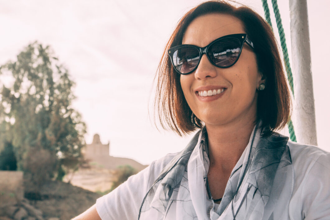 A woman with short brown hair smiles forcefully at the camera. She's wearing sunglasses and a white shirt and a light scarf around her neck.