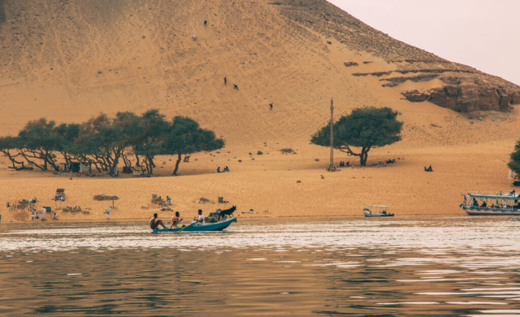 Nubian natives boat a small boat on the riverbank of the Nile river. The landscape of the riverbank is sandy with some trees and large hills.