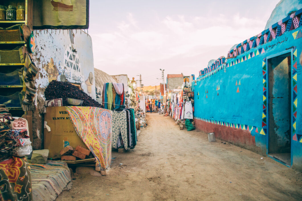 A street in the Nubian village lined with colorful street vendors selling fabrics, clothing, and gifts. On the righthand side of the photo is a stone building painted a bright aqua blue color, with colorful triangles painted across the side.