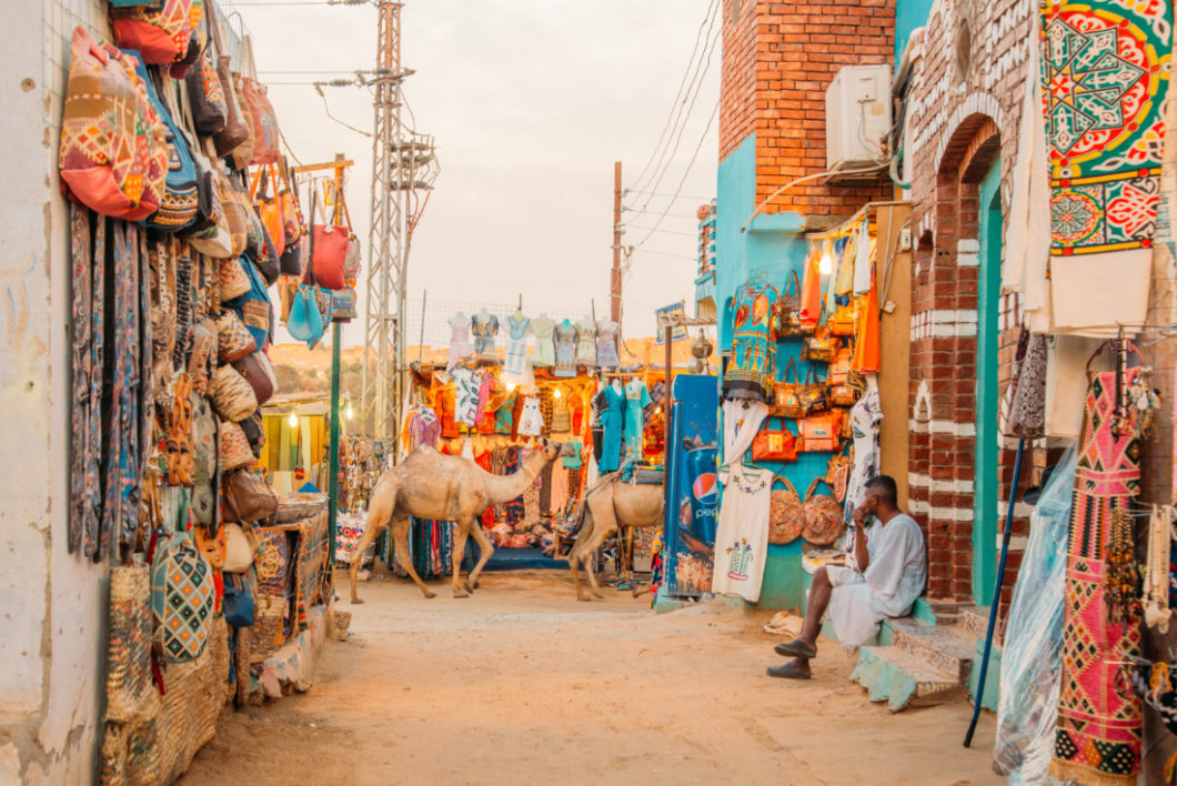 A street in the Nubian Village - camels walk along a dirt road that is lined with street vendors selling colorful gifts, fabrics, and clothing.