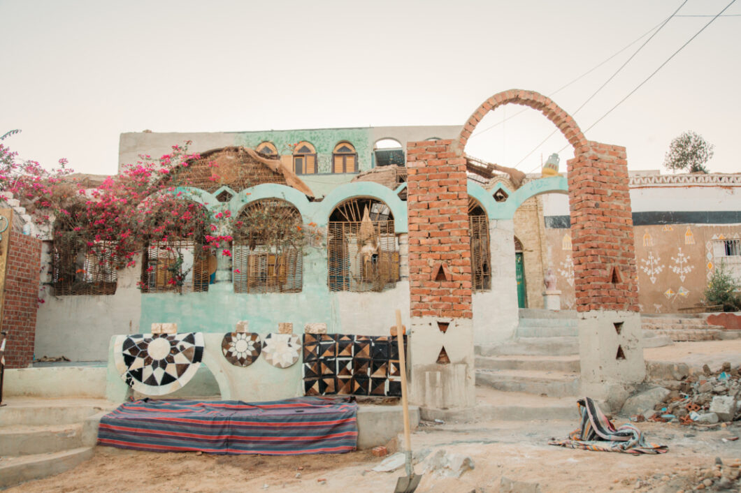 An image of the exterior façade of a home in the Nubian Village. The home has clay and stone walls and archways, some painted a light blue. Bushes of native flowers grow across the front of the building.