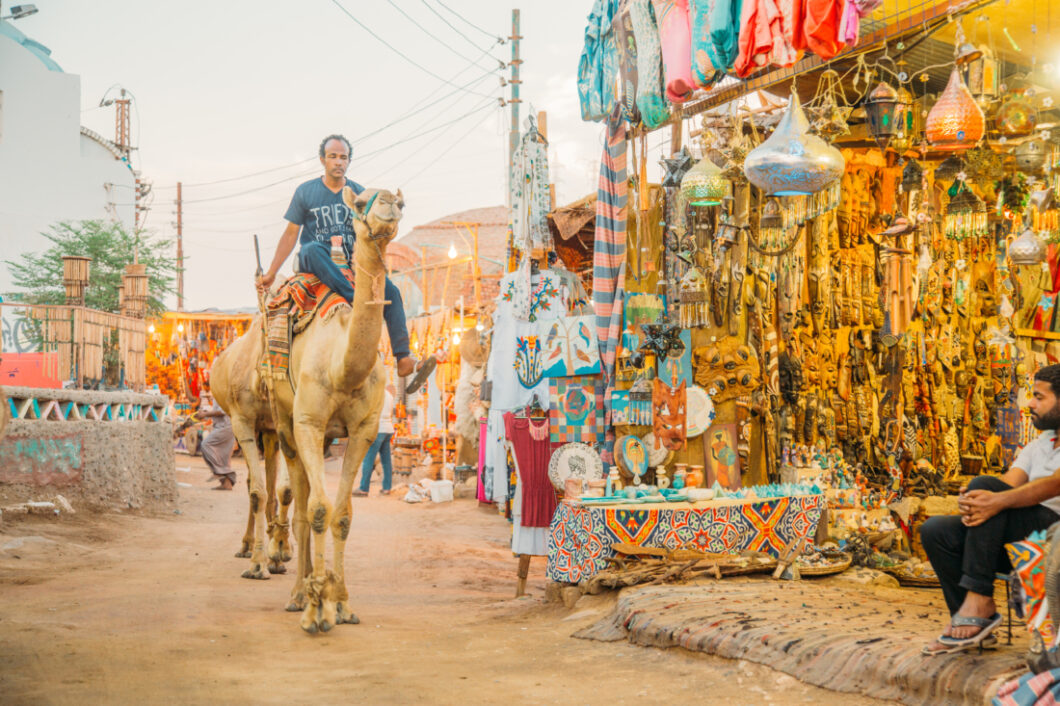 A man rides a camel along a dirt road in the center of the Nubian Village. He passes street vendor stands selling fabrics and jewelry. 