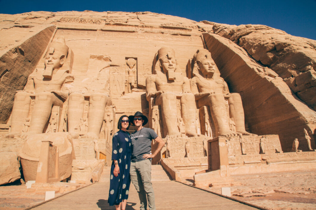 A man and woman pose in front of Abu Simbel, an ancient Egyptian temple with giant statues of pharaohs carved into a mountainside.