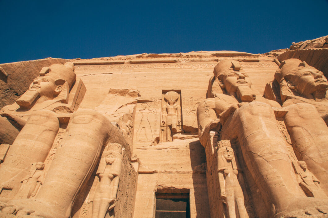 A photo shows the entrance to the Abu Simbel temple and the four giant stone Egyptian pharaoh statues that surround the entrance. The photo is at an upward angle, looking up at the giant statues and the sky.