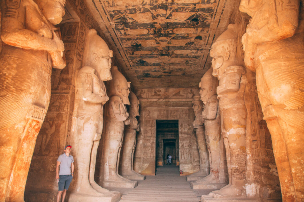 The cavernous entrance of the Ramses II temple in Abu Simbel. A long hall is lined with giant Egyptian statues, and the walls are covered floor to ceiling in artwork. A man stands next to one of the statues for scale.