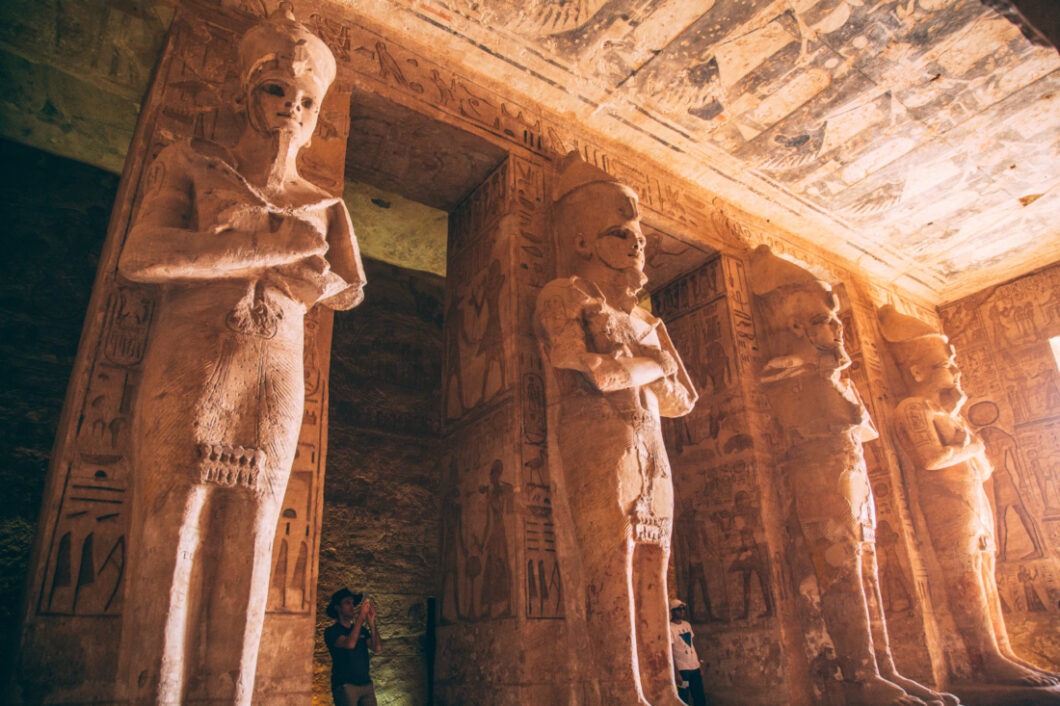 A photo inside one of the Abu Simbel temples. Four giant stone pillars rise to the ceiling, each with a different statue of Ramses II. The pillars, walls, and ceilings are filled with carvings. hieroglyphs, and artwork.
