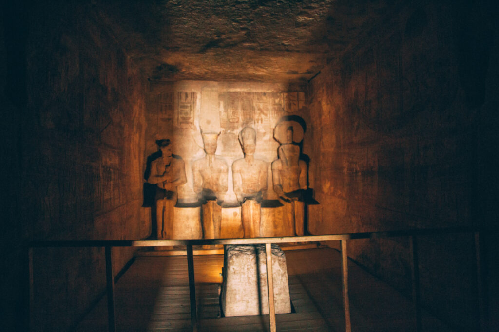 An interior wall inside a temple at Abu Simbel. The room is mostly dark, with a sliver of light falling on a display of statues and artwork against one wall.