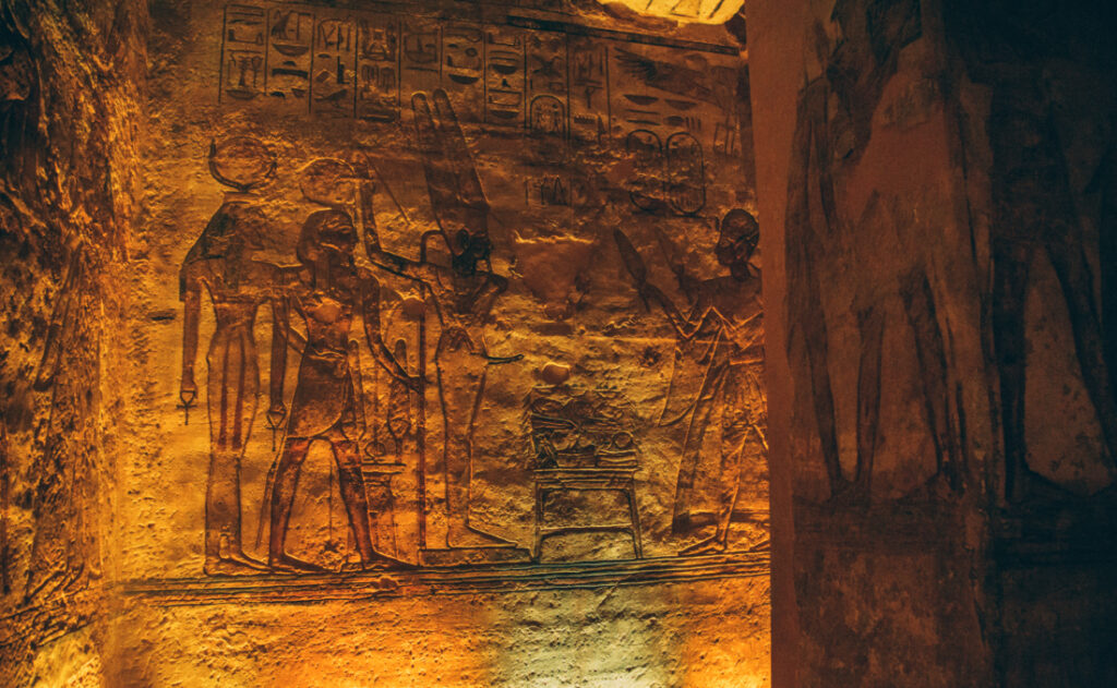 Dark, dimly list walls illuminated with orange and green light, showing ancient artwork carved onto the stone walls.