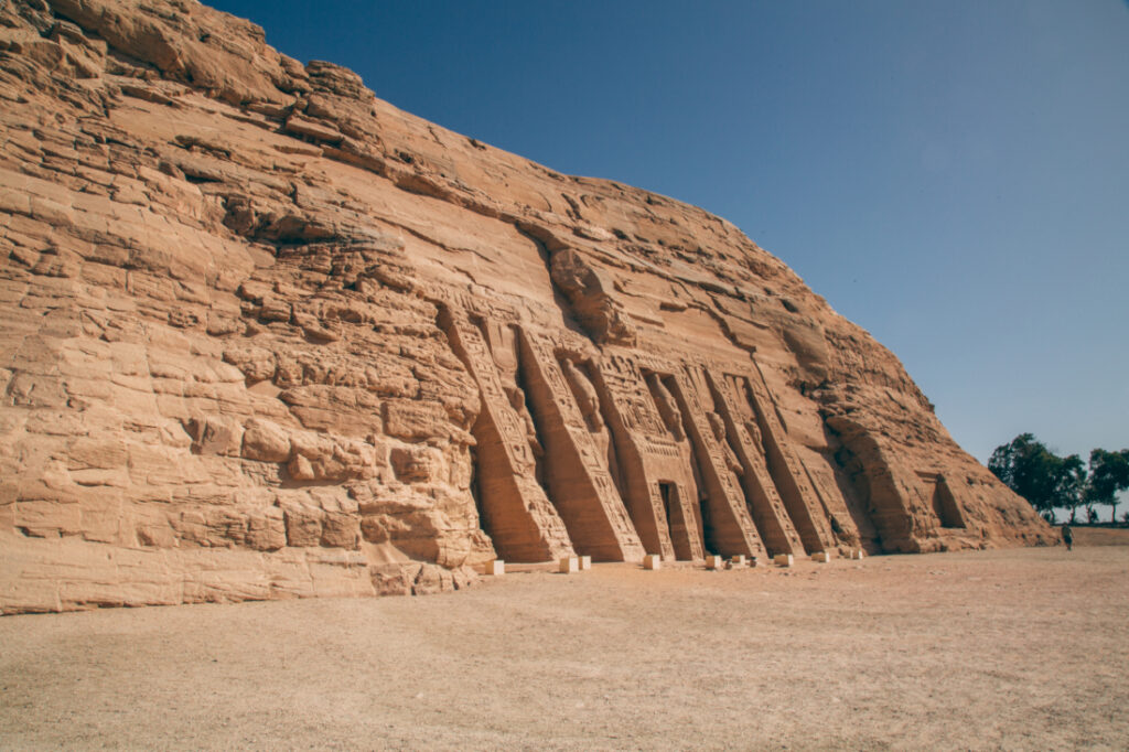 An exterior image of the outer façade of the Ramses II temple at Abu Simbel. The photo is taken at an angle from the left, showing the front of the temple and the clear blue sky in the background.