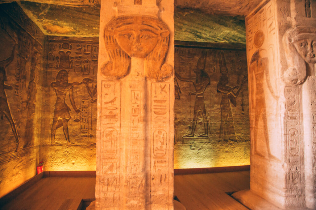 An interior image of a room inside a temple at Abu Simbel, showing a giant stone pillar and hallway. Every surface of the room is covered in carvings and artwork.