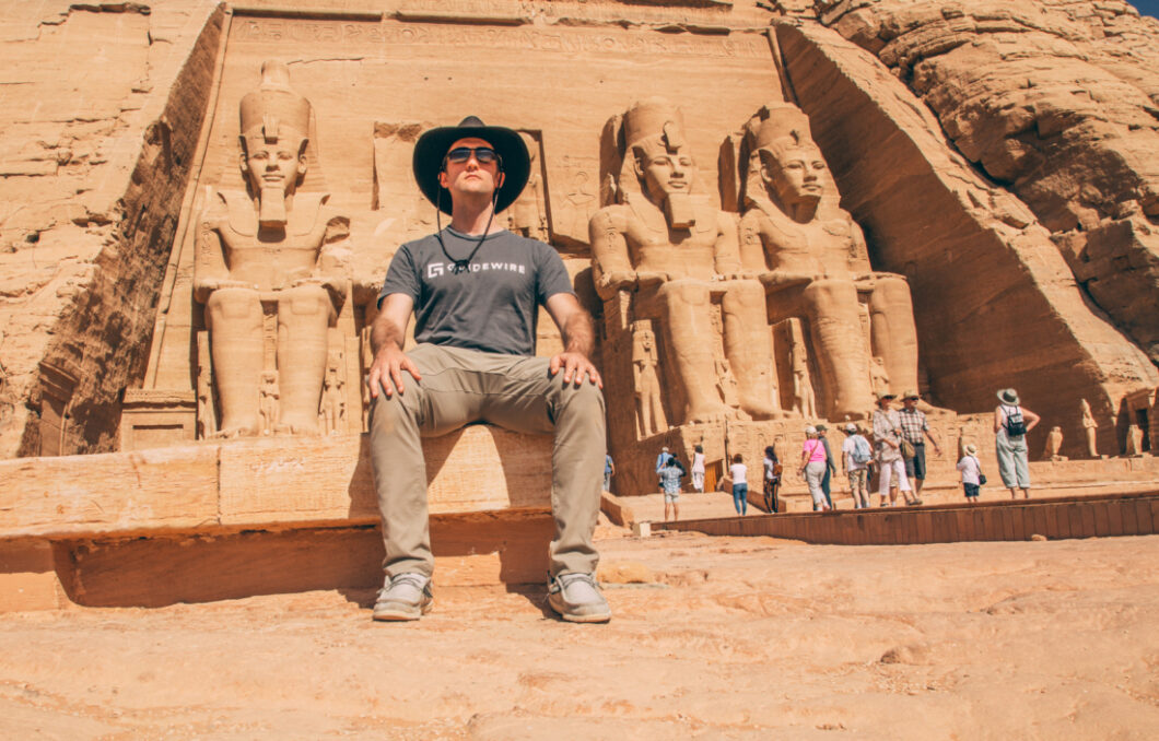 A man sits on a bench outside the front of an Abu Simbel temple. He's posing like one of the four Ramses statues that line the front of the temple. Tourists mull around in the background.