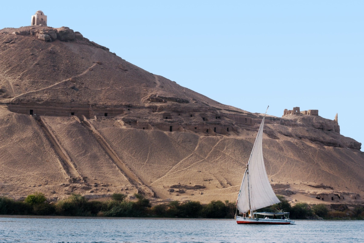 An Egyptian Felucca cruise boat sail over river Nile under Tombs of the Nobles in Aswan, Egypt.