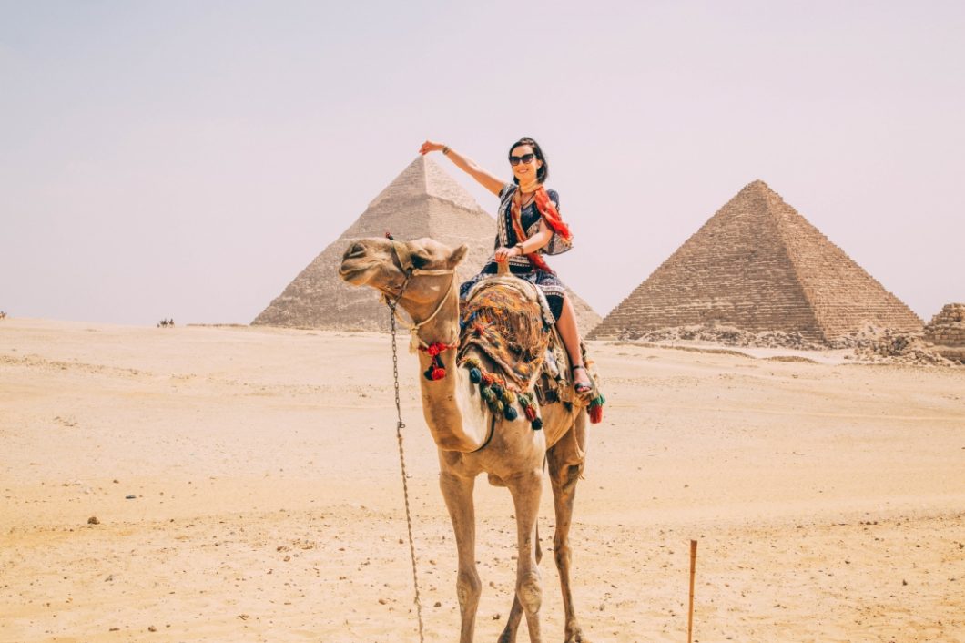 A woman riding a camel in front of the Pyramids of Giza wearing a dress.