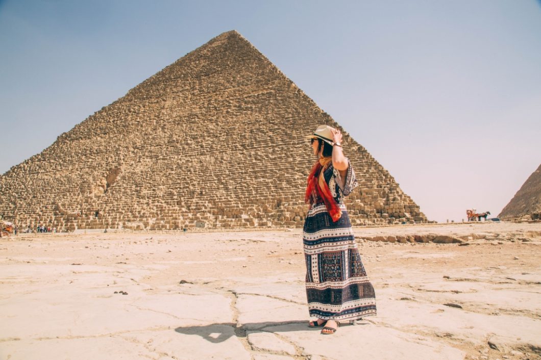 A woman is standing in front of the pyramids of Giza wearing a maxi dress, scarf, sandals, and hat.