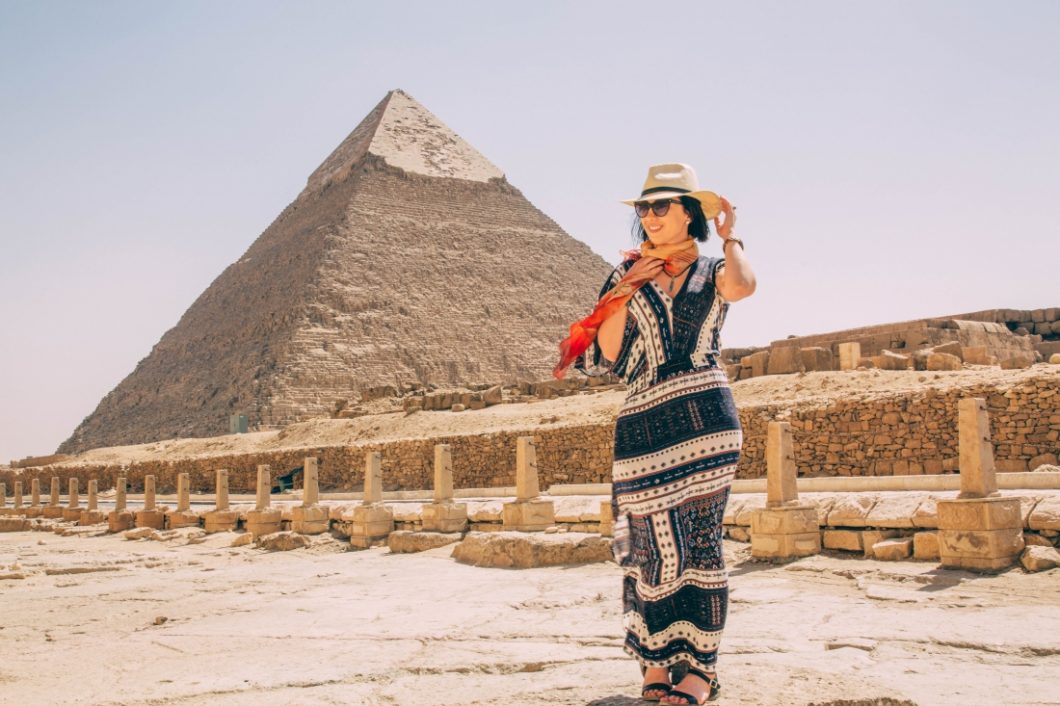 A woman standing in front of the pyramids in Giza, Egypt wearing a long dress, scarf, hat, and sunglasses.