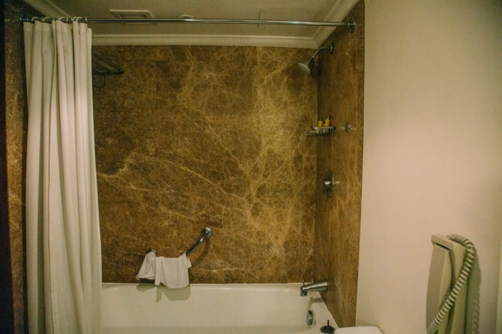 The shower in the bathroom of a suite at the Marriott Mena house is decently sized, with a white shower curtain and deep brown marble tiled walls.