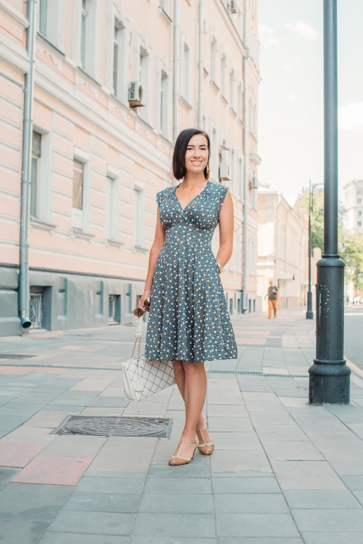 A young woman stands on a city sidewalk in the summertime. She's wearing a knee-length dress with a small all-over flower pattern. The dress has a v-neckline and pockets.