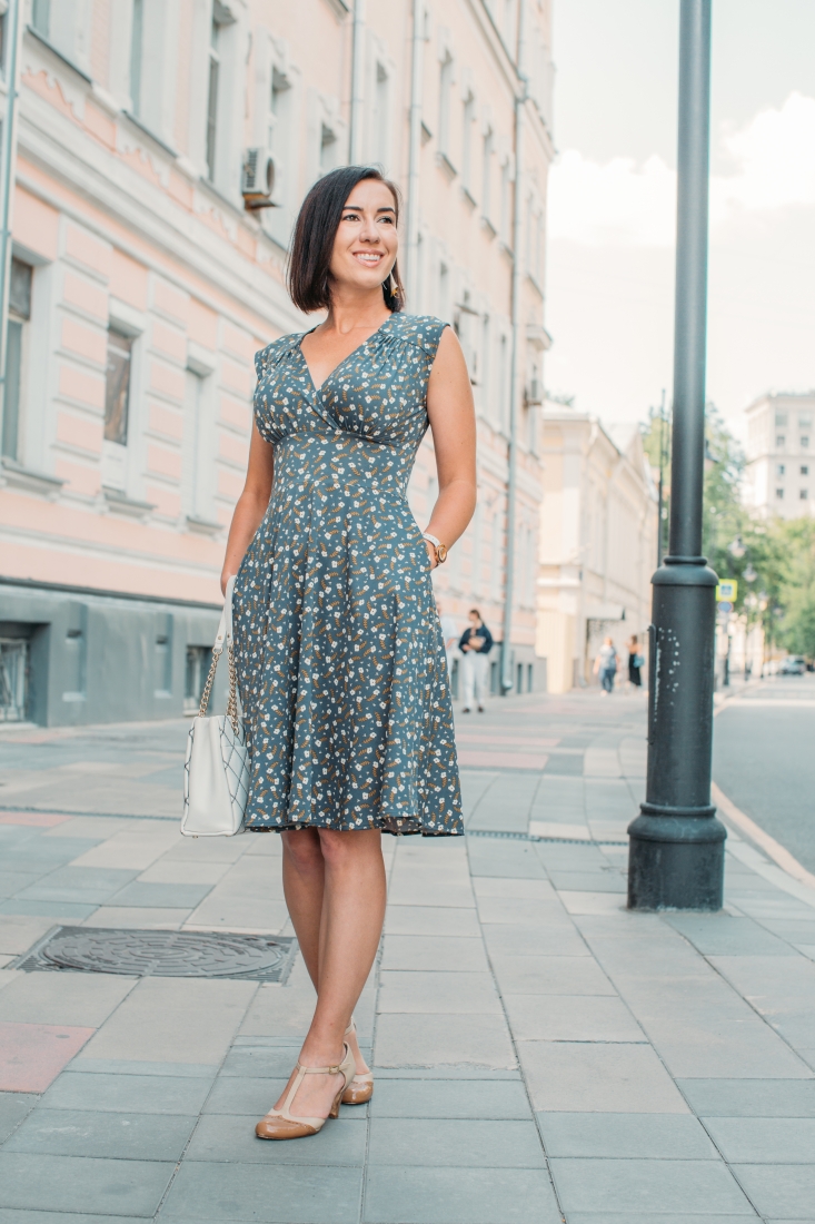 A woman poses on a city sidewalk wearing a stylish knee-length dress with an all-over clover flower pattern. The dress has a v-cut neckline and flowy skirt, and she pairs it with a white crosshatch patterned handbag and kitten heels.