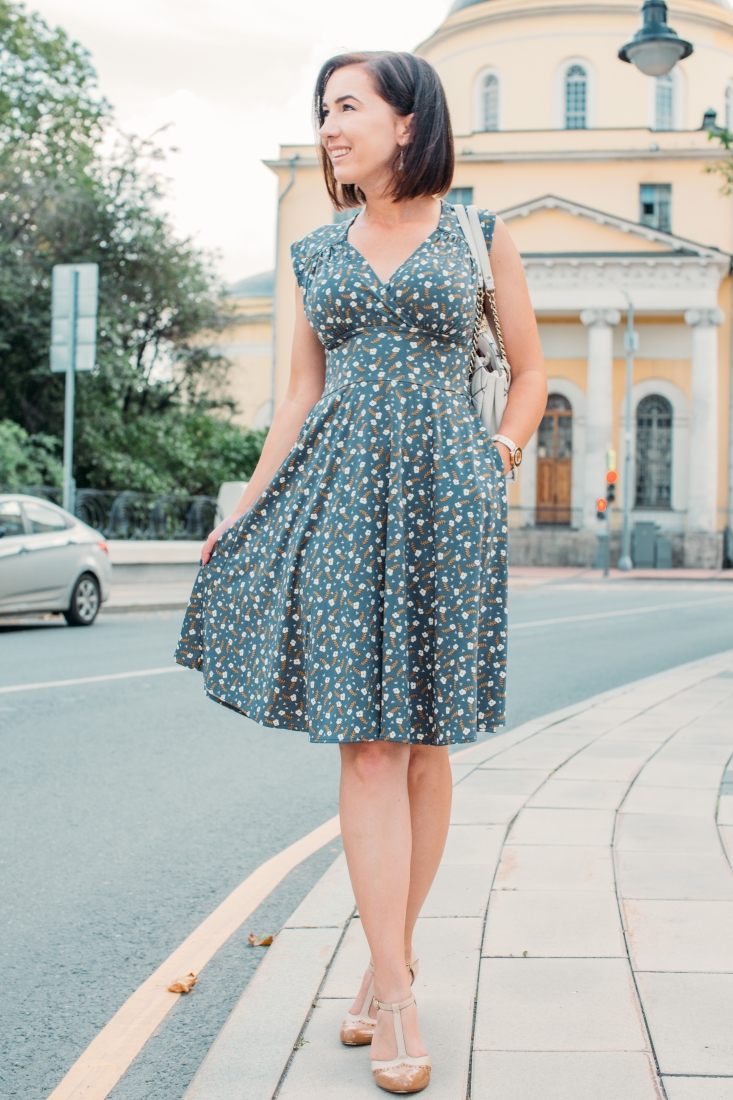 A woman poses on a city sidewalk, wearing a knee-length Karina dress in the Nora style. The dress is blue with an all over clover flower pattern, has a flowy skirt, and a v-cut neckline.