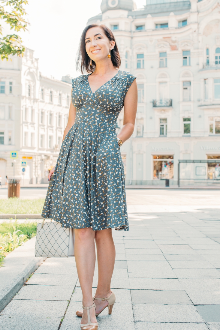 A woman poses on a city sidewalk wearing a knee-length dress with pockets. The flowy dress has a v-neckline and an all-over tiny flower pattern across the dress. She wears beige kitten heals and carries a tan crosshatch pattern bag.