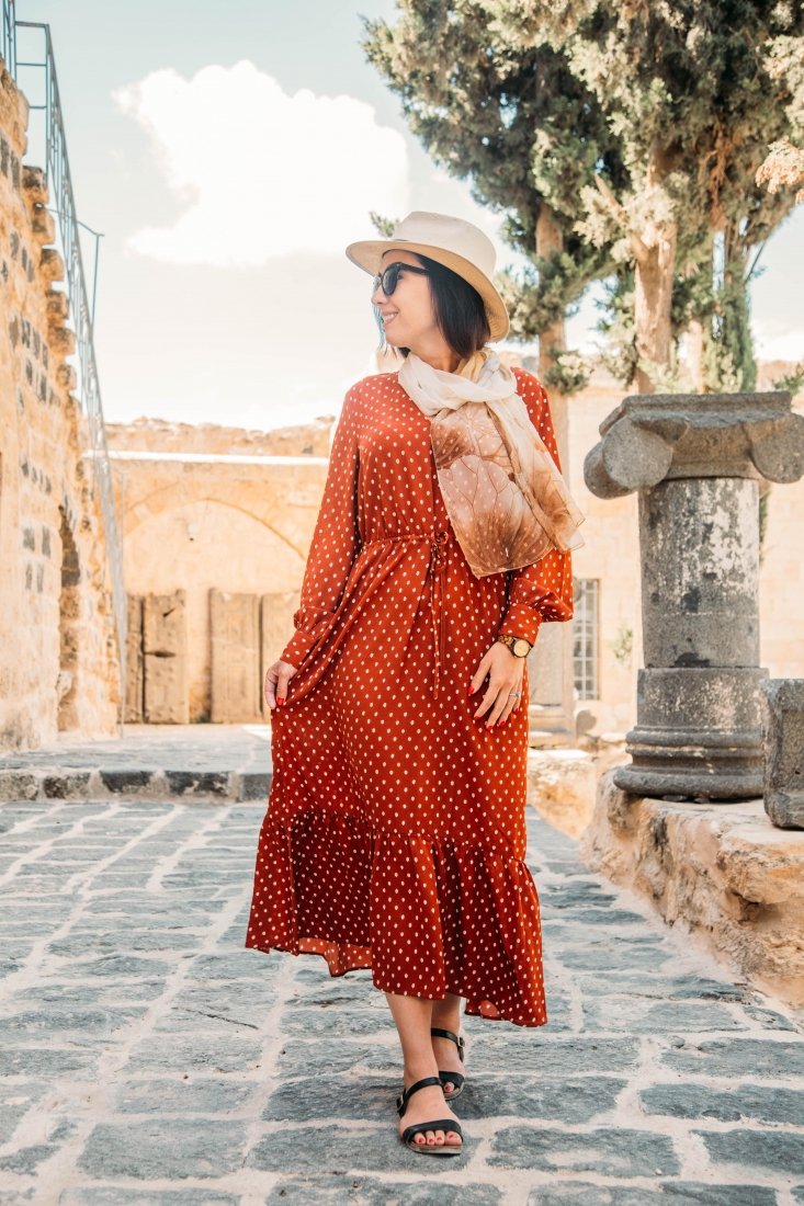 A woman walks down a cobblestone street, wearing a long-sleeved red maxi dress with small white polka dots, a sun hat, and tan scarf.