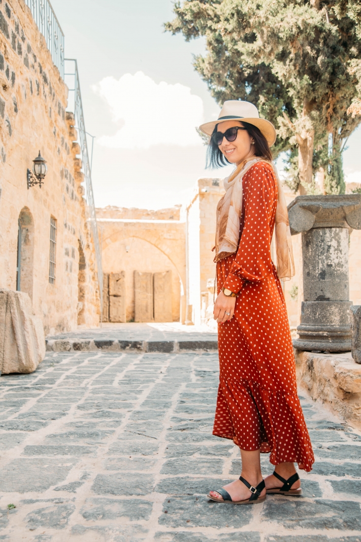 A woman poses on an empty cobblestone city street. She's wearing a long-sleeve maxi dress that is red with small white polka dots. The dress is accessorized with strappy sandals, a tan scarf and hat, and sunglasses.